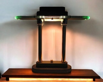 May include: A modern table lamp with a black rectangular shade and two vertical gold-colored supports. The lamp has a rectangular base and two horizontal arms with green accents. The lamp is placed on a wooden table.
