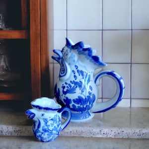 May include: Two blue and white ceramic rooster-shaped pitchers. The larger pitcher has a handle and a detailed floral design. The smaller pitcher has a similar design. Both are on a light-colored surface.