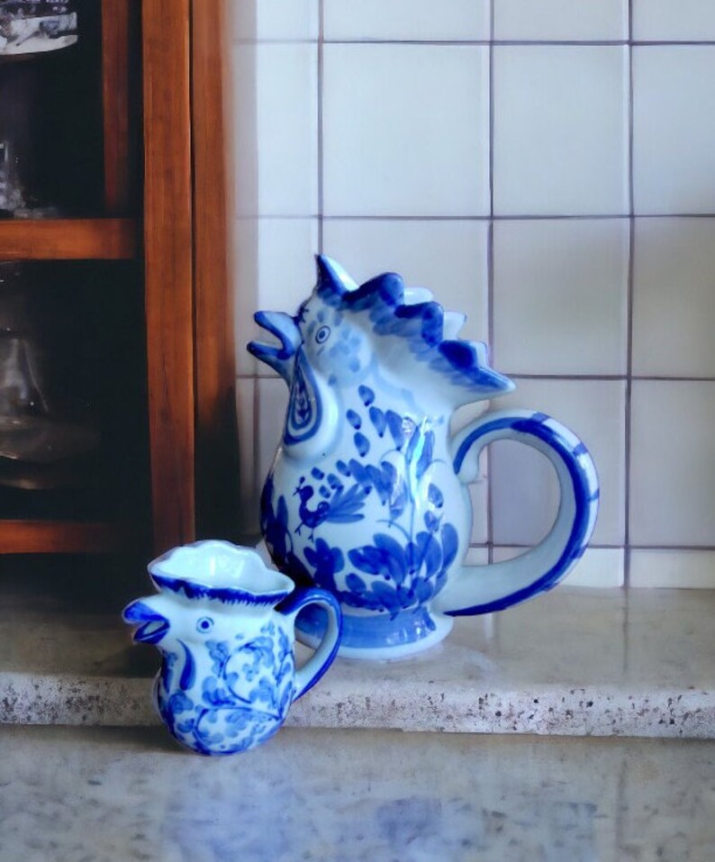 May include: Two blue and white ceramic rooster-shaped pitchers. The larger pitcher has a handle and a detailed floral design. The smaller pitcher has a similar design. Both are on a light-colored surface.