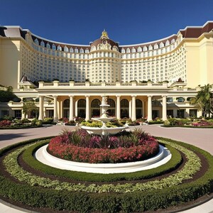 Puede incluir: Vista exterior de un gran hotel curvo con fachada amarillo claro y techo oscuro. Una fuente y parterres en primer plano, con palmeras y un cielo azul claro.