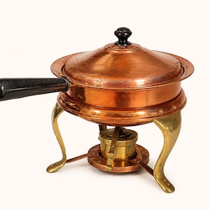May include: A copper chafing dish with a lid, featuring a long black handle and a black knob on top. The dish is supported by three curved brass legs and includes a central brass burner unit underneath for heating the contents of the pot.