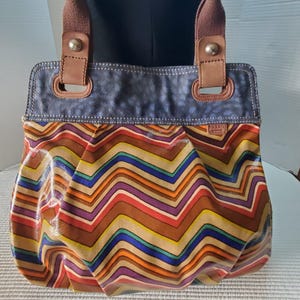 Peut inclure: Un sac cabas Fossil présentant un motif chevron multicolore vif dans des tons de bleu, violet, orange et beige. Le sac possède un panneau supérieur texturé bleu foncé, des attaches de sangle en cuir marron avec des rivets argentés et des anses en tissu marron.