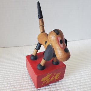 May include: A vintage wooden dog toy with a red base. The dog is tan with black spots, black ears, and a black tail. The base has the word "Fifi" in gold lettering. The toy is made of wood and has articulated joints.