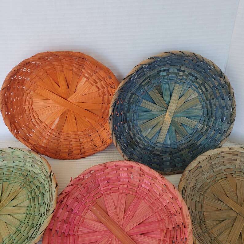 May include: A collection of colorful, round woven baskets in shades of orange, blue, pink, green, and tan. The baskets are made of woven material and have a circular design. The baskets are likely used for decorative or storage purposes.