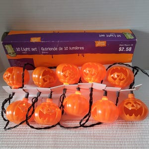 May include: A string of 10 orange plastic pumpkin-shaped lights with black wiring, presented in a purple and orange cardboard box labeled 10 Light set. The jack-o'-lanterns feature carved facial expressions and are arranged in two rows.