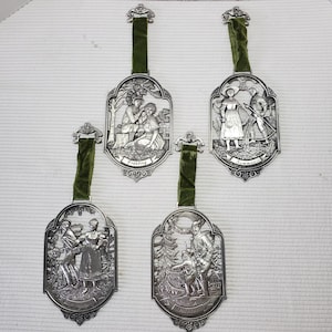 May include: Set of four silver-toned metal ornaments depicting scenes from the four seasons. Each ornament features a different scene with people and nature. The ornaments are hung on green ribbon.