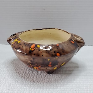 May include: A small, brown ceramic bowl with a speckled design in orange and yellow. The bowl has a rounded shape with a light beige interior and small feet. It features two small handles on either side.