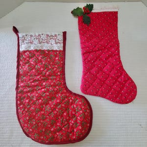 May include: Two red Christmas stockings. One has a white lace cuff and a pattern of green leaves and white flowers. The other has a pattern of white dots and is decorated with holly and berries.