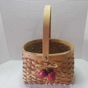 Peut inclure: Un panier en osier avec une poignée en bois et des accents décoratifs de pommes rouges. Le panier est fait d'un matériau tissé brun clair et comporte une poignée fixée avec une attache métallique. Le panier est décoré d'un nœud en raphia et de deux ornements de pommes rouges.