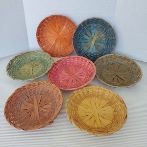 May include: A collection of seven woven baskets in various colors, including orange, blue, green, pink, brown, and yellow. The baskets are round and oval-shaped, with a woven design. The baskets are made of a natural material, possibly bamboo or wicker.