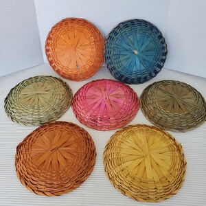 May include: Seven round woven baskets in various colors, including orange, blue, pink, and yellow. The baskets are made of thin, natural-colored strips of material, arranged in a radial pattern. The baskets are likely decorative or for storage.