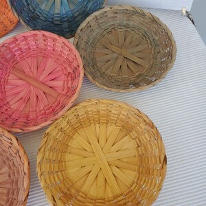 May include: A collection of round, woven baskets in various colors, including pink, blue, and yellow. The baskets are made of natural materials and have a radial pattern in the center.