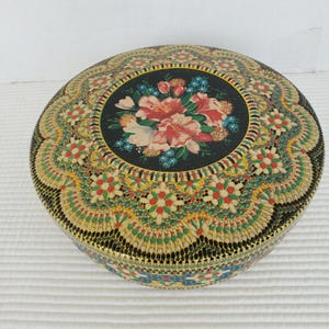 May include: Circular vintage metal tin with a floral design on the lid. The lid has a black centre with pink and blue flowers. The tin's border has a detailed, colourful pattern with red, yellow, and green accents. The sides are black with a gold trim.