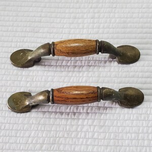 May include: A pair of vintage cabinet handles. Each handle has a central wooden section with a warm brown finish, set between aged metal hardware. The metal has a tarnished, antique look. The handles are designed for easy grip and installation.