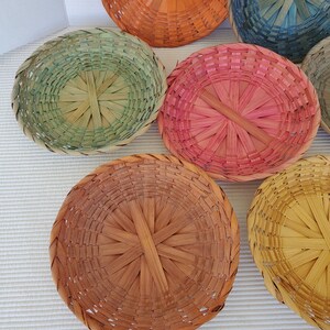 May include: A collection of colorful, round woven baskets in shades of pink, green, orange, blue, and yellow. The baskets are made of thin, natural-looking material and have a simple, rustic design. The baskets are arranged on a white surface.