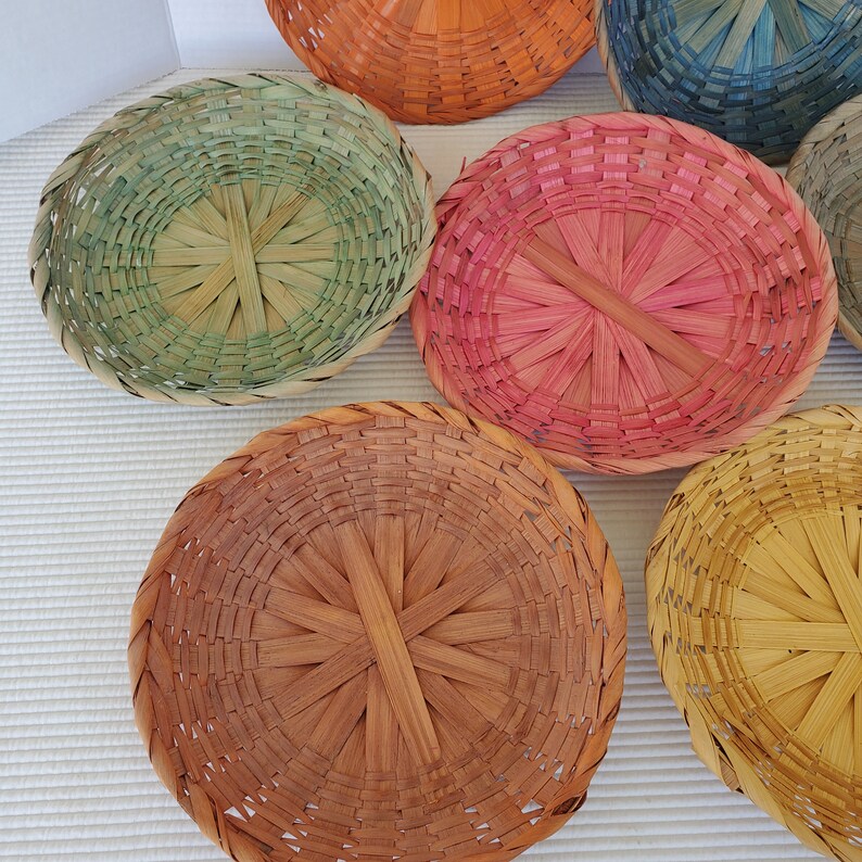 May include: A collection of colorful, round woven baskets in shades of pink, green, orange, blue, and yellow. The baskets are made of thin, natural-looking material and have a simple, rustic design. The baskets are arranged on a white surface.
