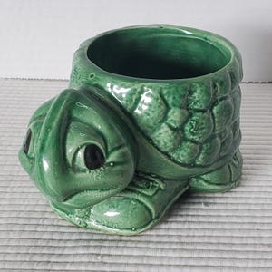 Peut inclure: Un pot de fleur en céramique verte en forme de tortue avec une large ouverture. La tortue a une carapace détaillée et un visage souriant avec des yeux noirs. Le pot est conçu pour contenir de petites plantes ou des fleurs.