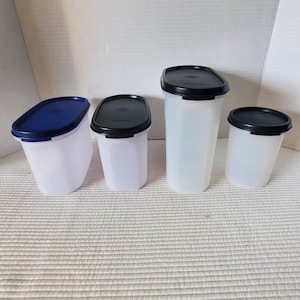 May include: A set of four translucent plastic food storage containers with lids. Three containers feature black oval lids, while one container has a blue oval lid. The containers vary in height and width, with a tall rectangular shape and rounded edges.