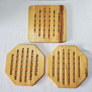 May include: Three light brown wooden trivets with a natural grain pattern. One trivet is square with rounded corners, and two are octagonal. Each piece features a series of vertical cutouts and small circular holes arranged in a grid pattern.