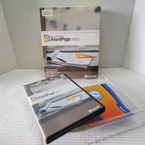 May include: Microsoft FrontPage 2003 software package. The box and CD case feature the FrontPage 2003 logo and product name. The box shows a diagram and the CD case is black with a silver label. A clear plastic tray holds additional inserts.