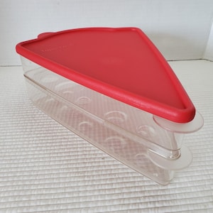 May include: A triangular food storage container with a clear base and a solid red lid. The container features a wedge-shaped design with rounded corners and a series of circular indentations along the sides. The word Tupperware is embossed on the lid.