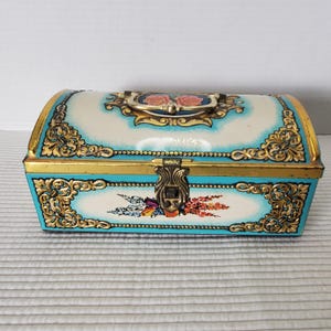 May include: Vintage decorative chest with a curved top. The chest is painted in a light blue and white color scheme, with gold trim and ornate floral designs. A handle and clasp are visible on the top and front.