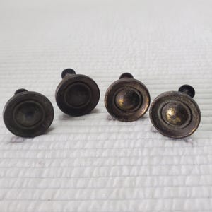 May include: Four antique brass cabinet knobs. Each knob has a circular design with a raised centre and a small, rounded stem. The knobs have a tarnished, aged appearance, suggesting they are vintage or vintage-inspired.