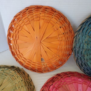 May include: Overhead view of several round woven baskets in various colors. The largest basket is orange, with a woven pattern radiating from the center. Other baskets are visible in shades of green, blue, and pink. The baskets are made of thin, woven strips of material.