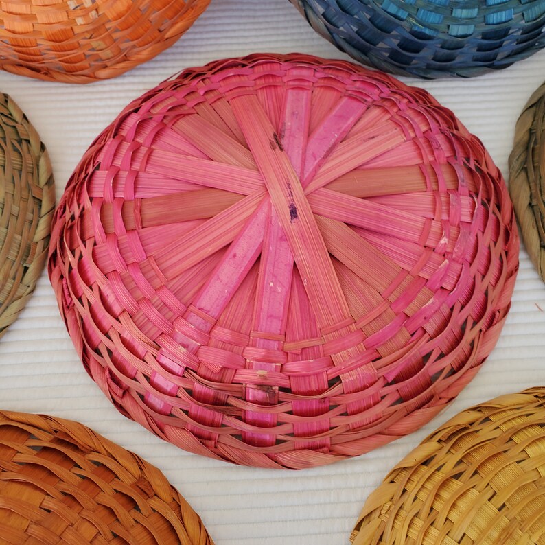 May include: A collection of colorful, woven baskets in various shades, including pink, orange, and blue. The baskets are round with a woven pattern, showcasing the craftsmanship of the design. The image highlights the texture and vibrant colors of the handmade items.