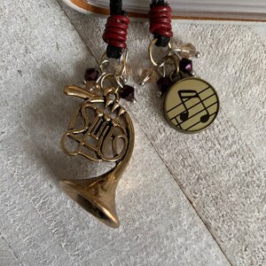 May include: A gold-colored french horn charm with a music note charm on a red and black cord necklace. The charms are accented with small, clear and burgundy glass beads.