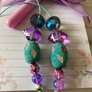 May include: Two teal and silver patterned beads with purple and blue glass beads hanging from a light blue cord.