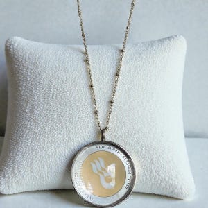 May include: Silver-toned necklace with a round pendant. The pendant features a cream-colored background with a white logo and the text "REGAL PRINCESS - MAR 17, 2019" and "DYLAN HARRIS". The necklace is displayed on a white pillow.