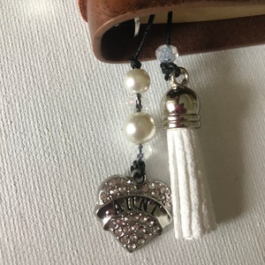 May include: A silver heart-shaped charm with the word "AUNT" in rhinestones hangs from a black string with white pearl beads and a white tassel.
