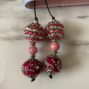 May include: Two pink and silver beaded bookmarks with a black cord. The top bead is covered in clear and pink rhinestones. The bottom bead is a dark pink with silver accents.