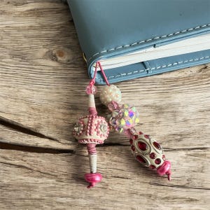 May include: A pink and white beaded bookmark with a light blue book. The bookmark features various decorative beads, including a large, embellished oval bead with red accents and a smaller, round bead with pink and white details. The bookmark is attached to the book with a pink string.