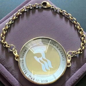 May include: Gold-toned bracelet with a circular pendant. The pendant features the words "REGAL PRINCESS - MAR 17 2018" and "DYLAN BARNES" around a logo. The bracelet is on a purple surface.