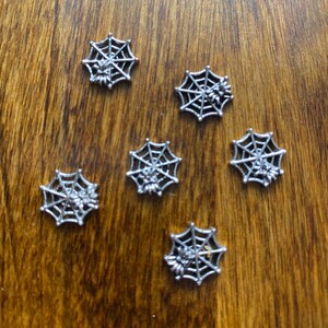 Op de afbeelding: Zes zilveren spinnenweb-bedels met een spin in het midden van elk web. De bedels liggen op een houten oppervlak.