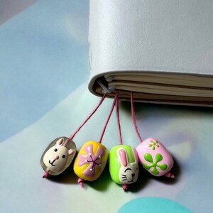 May include: A set of five colorful bookmarks with different animal and floral designs. The bookmarks are attached to a white notebook with pink strings.