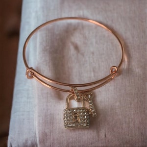 May include: A gold bangle bracelet with a padlock and key charm. The padlock and key are both covered in clear rhinestones.