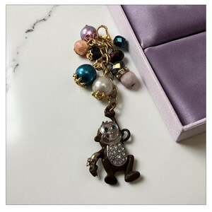 May include: A charm featuring a brown monkey pendant with a rhinestone-studded belly, hanging from a gold chain with colorful beads. The beads include teal, pearl, and pink tones. The charm is next to a purple box.