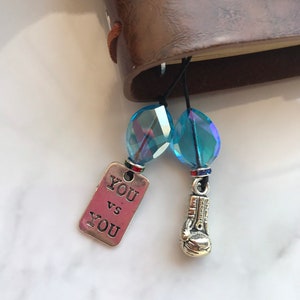 May include: A silver charm with the text "YOU VS YOU" and a silver boxing glove charm attached to a black cord with two blue crystal beads.