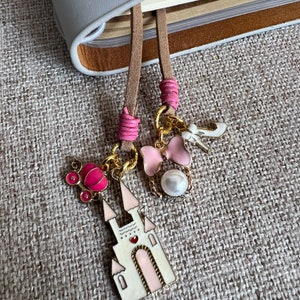 Könnte beinhalten: Ein rosa-weißer Schloss-Anhänger mit einer rosa Kutsche, einer rosa Schleife und weißen Perlenakzenten. Der Anhänger ist an einem braunen Leder-Lesezeichen befestigt.