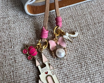 Könnte beinhalten: Ein rosa-weißer Schloss-Anhänger mit einer rosa Kutsche, einer rosa Schleife und weißen Perlenakzenten. Der Anhänger ist an einem braunen Leder-Lesezeichen befestigt.
