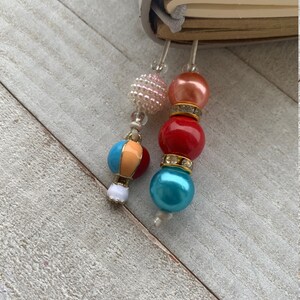 May include: Two colorful beaded bookmarks with a white, orange, and blue bead, a red bead with rhinestones, a turquoise bead, and a pink bead with rhinestones.
