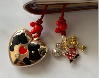 Könnte beinhalten: Ein goldener herzförmiger Anhänger mit einem schwarz-weißen Design und einem roten Herzen in der Mitte. Der Anhänger ist an einer roten Schnur mit roten Perlen befestigt. Ein goldener Mickey-Maus-Anhänger mit roten und weißen Punkten und ein goldener Schlüsselanhänger sind ebenfalls an der Schnur befestigt.