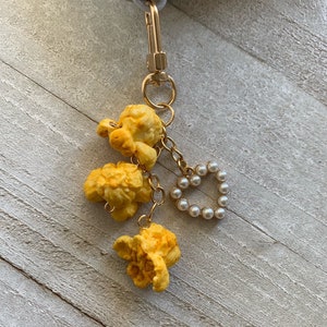 Puede incluir: Un llavero dorado con tres dijes de palomitas de maíz amarillas y un dije de corazón de perlas.