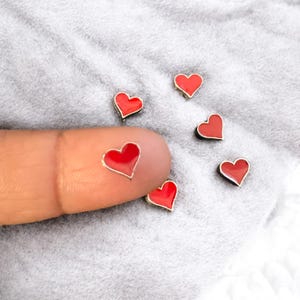 Op de afbeelding: Zes kleine rode hartvormige bedels met een zilveren rand. De bedels zijn verspreid over een wit, pluche oppervlak. Een bedel wordt vastgehouden door een vinger.