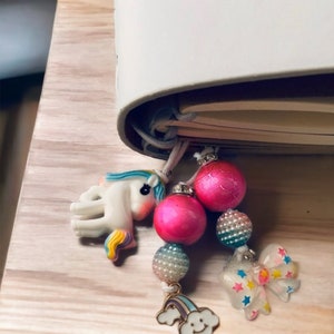 May include: A white notebook with a unicorn, rainbow, cloud, and star charm attached to the spine. The charms are made of plastic and have a colorful design.