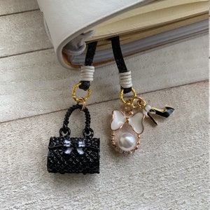 Könnte beinhalten: Zwei schwarze und goldene Handtaschen-Anhänger mit weißen Akzenten hängen an einer schwarzen Kordel. Ein Anhänger ist eine kleine schwarze Handtasche mit einer Schleife. Der andere Anhänger zeigt eine weiße Schleife, eine Perle und einen schwarzen High Heel.