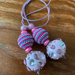 May include: A purple elastic hair tie with two large beads. One bead is pink, blue, and white striped. The other bead is light purple with clear and pink rhinestones.